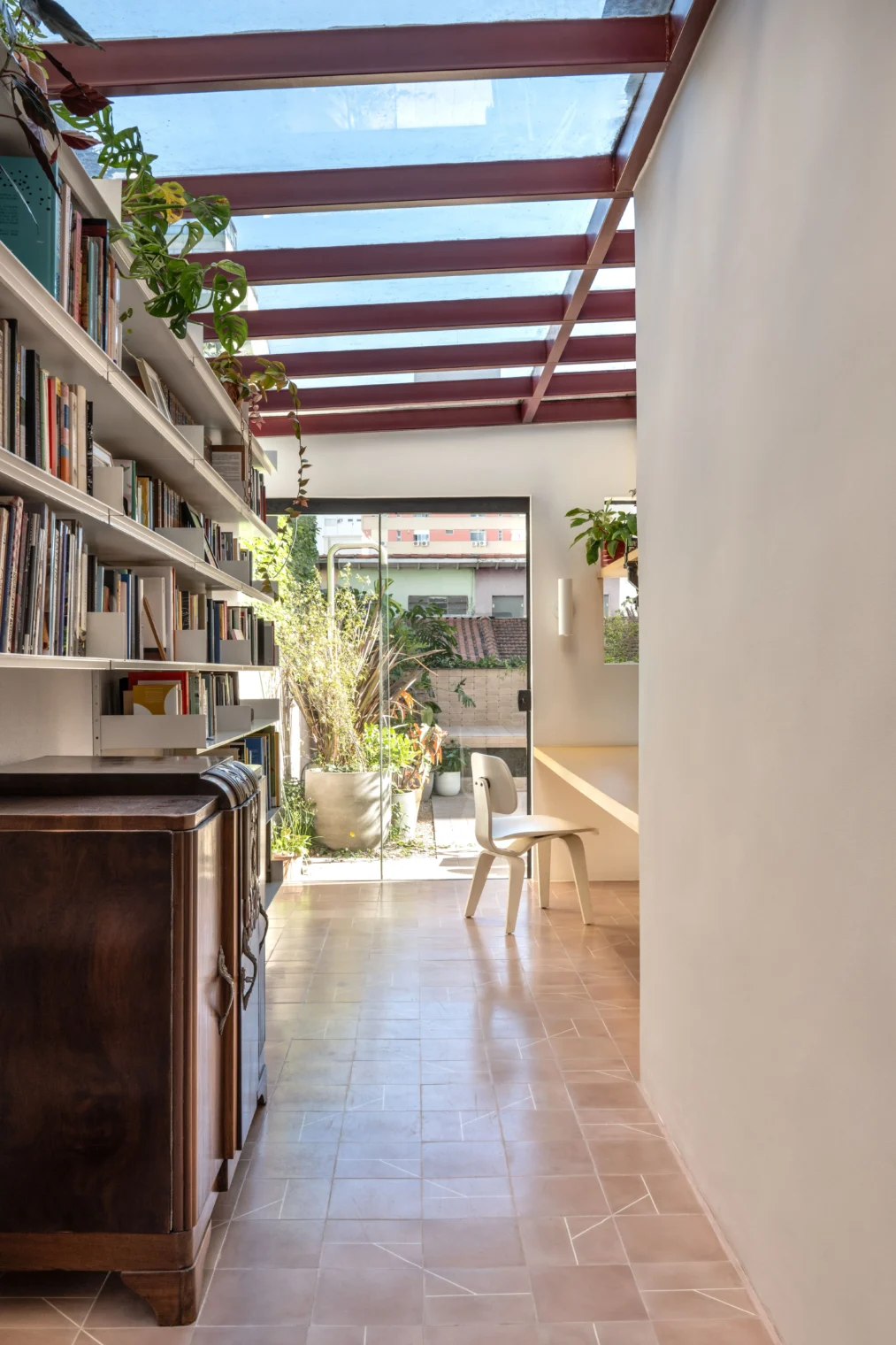 Projeto de corredor com função de home office e biblioteca, apresentando cobertura de vidro com estrutura metálica bordô, estante linear de livros, piso em ladrilho hidráulico rosado e porta de vidro para o quintal.