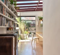 Projeto de corredor com função de home office e biblioteca, apresentando cobertura de vidro com estrutura metálica bordô, estante linear de livros, piso em ladrilho hidráulico rosado e porta de vidro para o quintal.