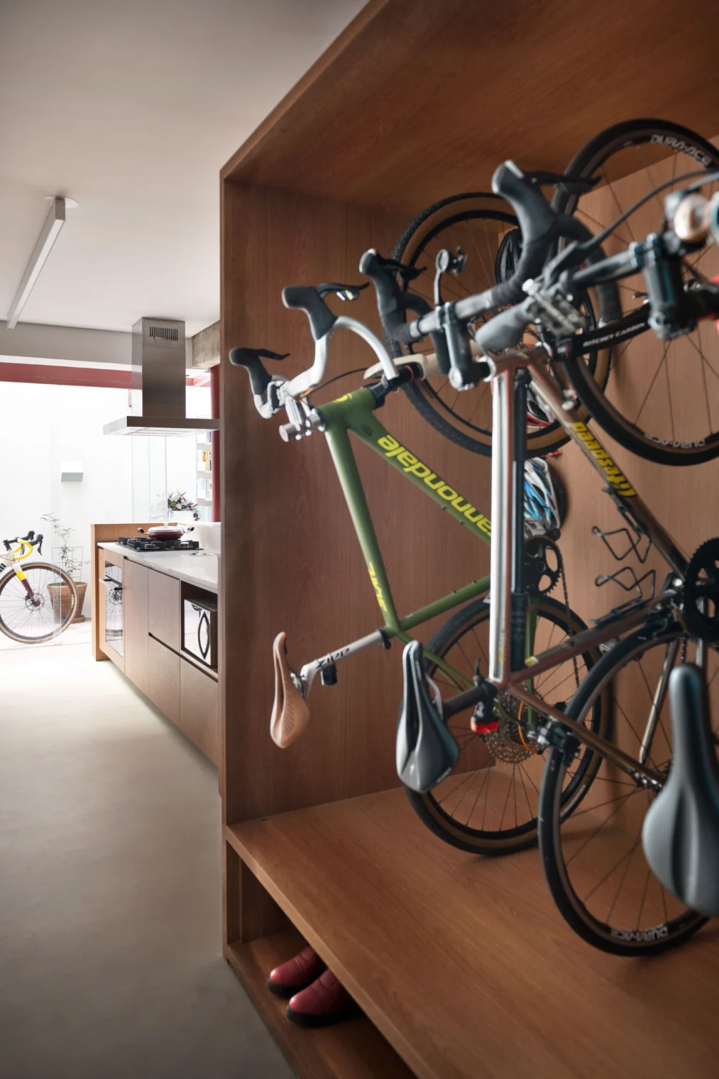 Projeto de interiores com bicicletário vertical desenhado em nicho de marcenaria de madeira. Ao fundo, vista para uma cozinha integrada com armários de madeira, piso claro e coifa de inox.