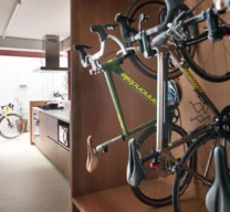 Projeto de interiores com bicicletário vertical desenhado em nicho de marcenaria de madeira. Ao fundo, vista para uma cozinha integrada com armários de madeira, piso claro e coifa de inox.