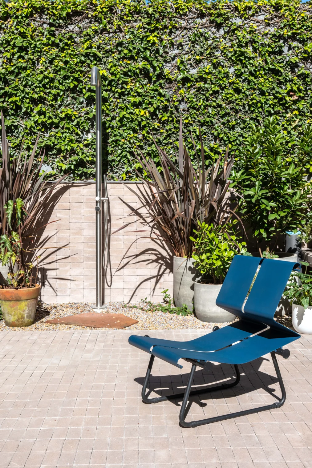 Projeto de área externa com chuveirão de piso em aço inox, jardim com parede verde, piso claro de tijolinho e uma cadeira de azul assinada por Paulo Mendes da Rocha.