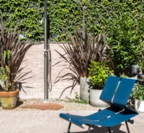 Projeto de área externa com chuveirão de piso em aço inox, jardim com parede verde, piso claro de tijolinho e uma cadeira de azul assinada por Paulo Mendes da Rocha.