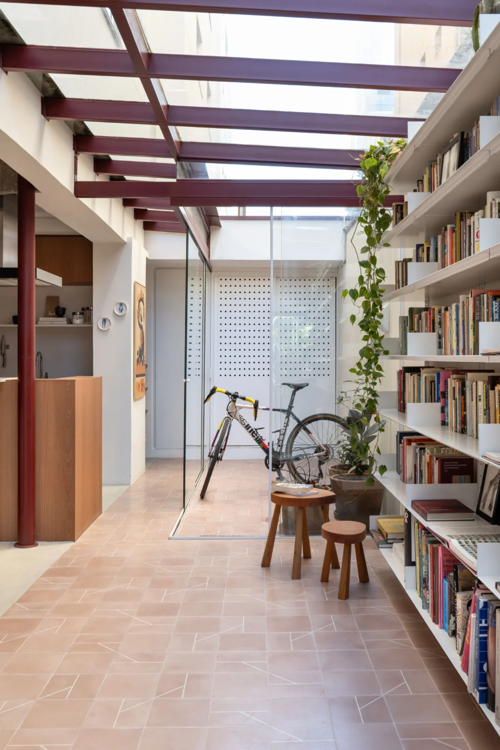 Projeto de corredor iluminado por teto de vidro com estrutura metálica bordô, piso em ladrilho geométrico rosa, grande estante de livros branca e vista para um solário fechado em vidro.