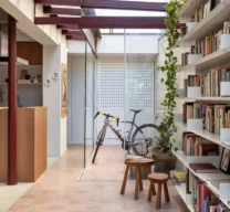 Projeto de corredor iluminado por teto de vidro com estrutura metálica bordô, piso em ladrilho geométrico rosa, grande estante de livros branca e vista para um solário fechado em vidro.
