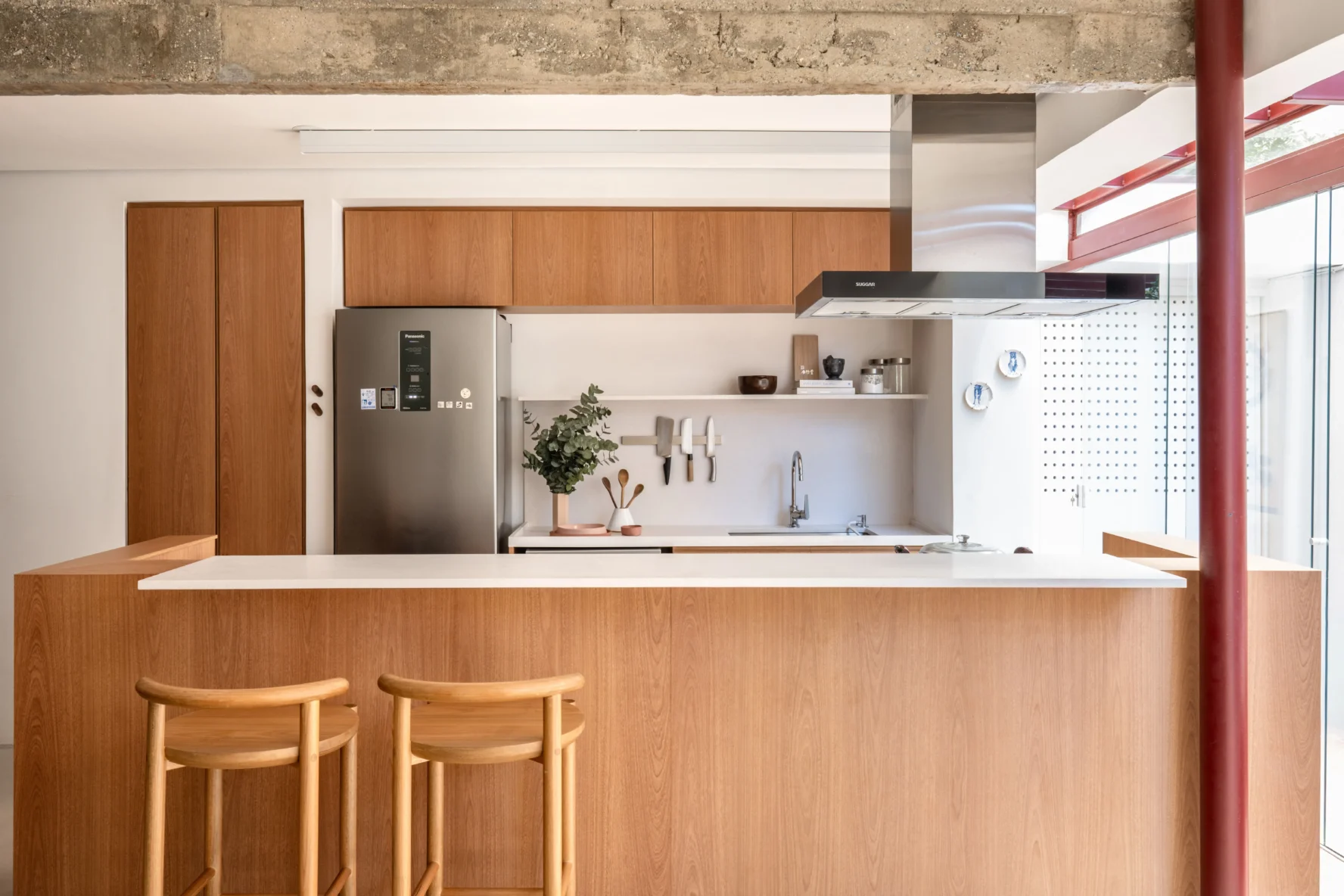 Projeto de cozinha com ilha central em madeira de tom médio, bancada branca, banquetas de madeira, eletrodomésticos em inox e viga de concreto aparente no teto.