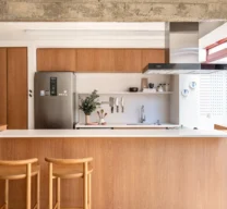Projeto de cozinha com ilha central em madeira de tom médio, bancada branca, banquetas de madeira, eletrodomésticos em inox e viga de concreto aparente no teto.