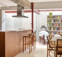 Texto Alternativo: Projeto de cozinha e sala de jantar integradas com ilha de madeira, vigas de concreto aparente no teto, piso monolítico claro e mesa com cadeiras de palhinha.