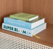 Detalhe de estante de madeira com porta em palhinha clara e três livros de arte e botânica empilhados sobre a bancada.