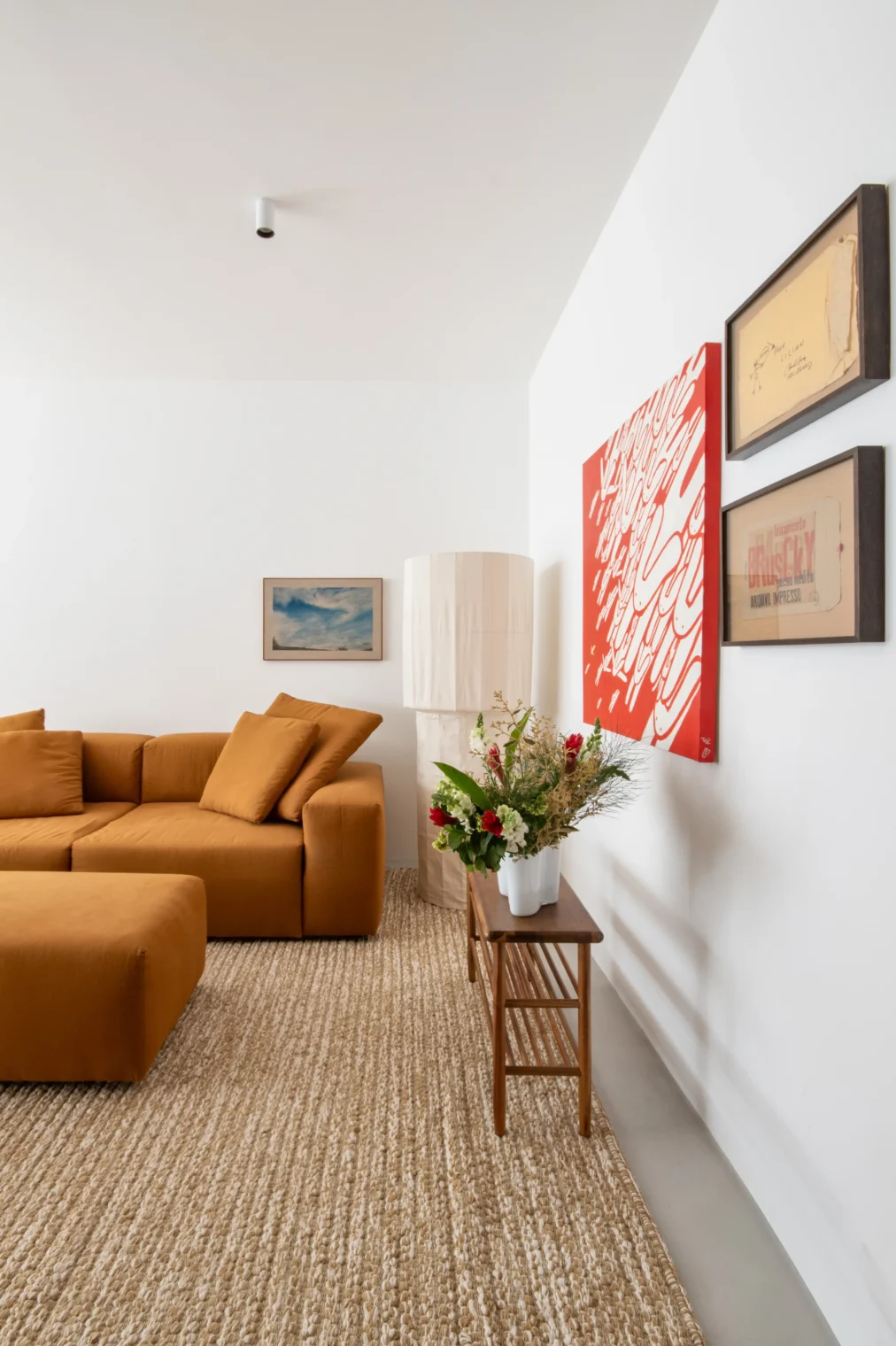 Projeto de sala de estar com sofá modular em tom terracota, tapete texturizado de fibra natural, banco de apoio em madeira e composição de quadros com destaque para uma tela vermelha.