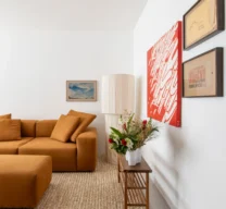 Projeto de sala de estar com sofá modular em tom terracota, tapete texturizado de fibra natural, banco de apoio em madeira e composição de quadros com destaque para uma tela vermelha.