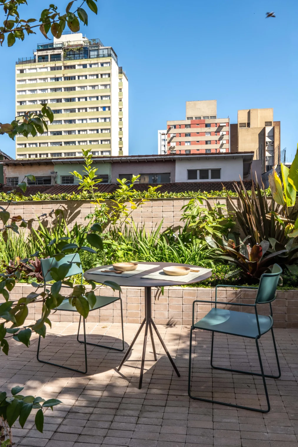 Projeto de área externa com mesa redonda e cadeiras metálicas verdes sobre piso de lajota tipo tijolinho, ladeados por canteiro elevado com paisagismo tropical e vista para a cidade.