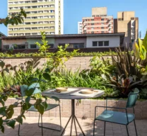Projeto de área externa com mesa redonda e cadeiras metálicas verdes sobre piso de lajota tipo tijolinho, ladeados por canteiro elevado com paisagismo tropical e vista para a cidade.