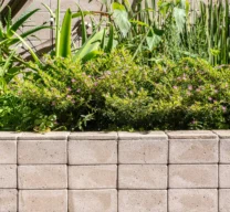 Detalhe de canteiro elevado e muro de fundo revestidos em lajotas cerâmicas tipo tijolinho, abrigando paisagismo com folhagens verdes e pequenas flores.