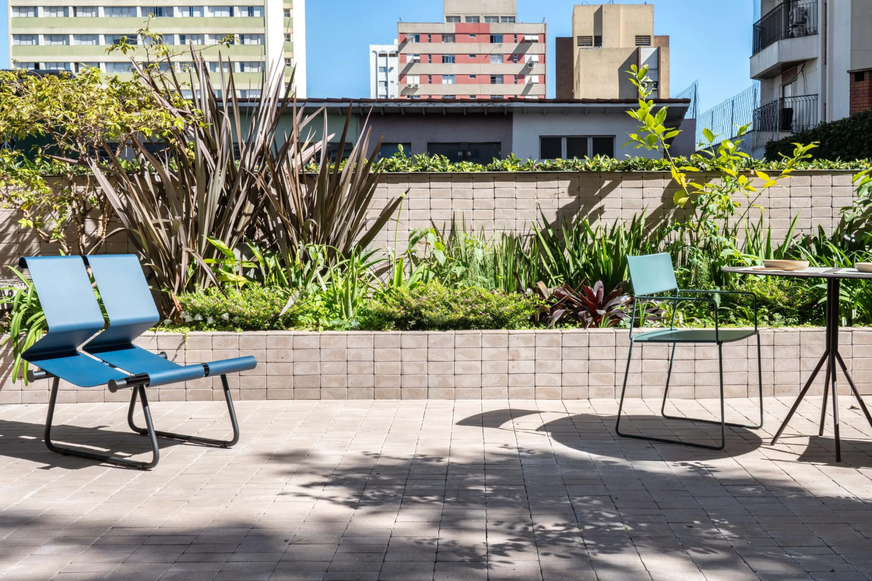Projeto de área externa com piso e canteiro elevado revestidos em lajota cerâmica tipo tijolinho. O espaço conta com uma cadeiras lounge azul, uma mesa redonda com cadeira verde e paisagismo tropical, tendo prédios da cidade ao fundo.