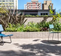 Projeto de área externa com piso e canteiro elevado revestidos em lajota cerâmica tipo tijolinho. O espaço conta com uma cadeiras lounge azul, uma mesa redonda com cadeira verde e paisagismo tropical, tendo prédios da cidade ao fundo.