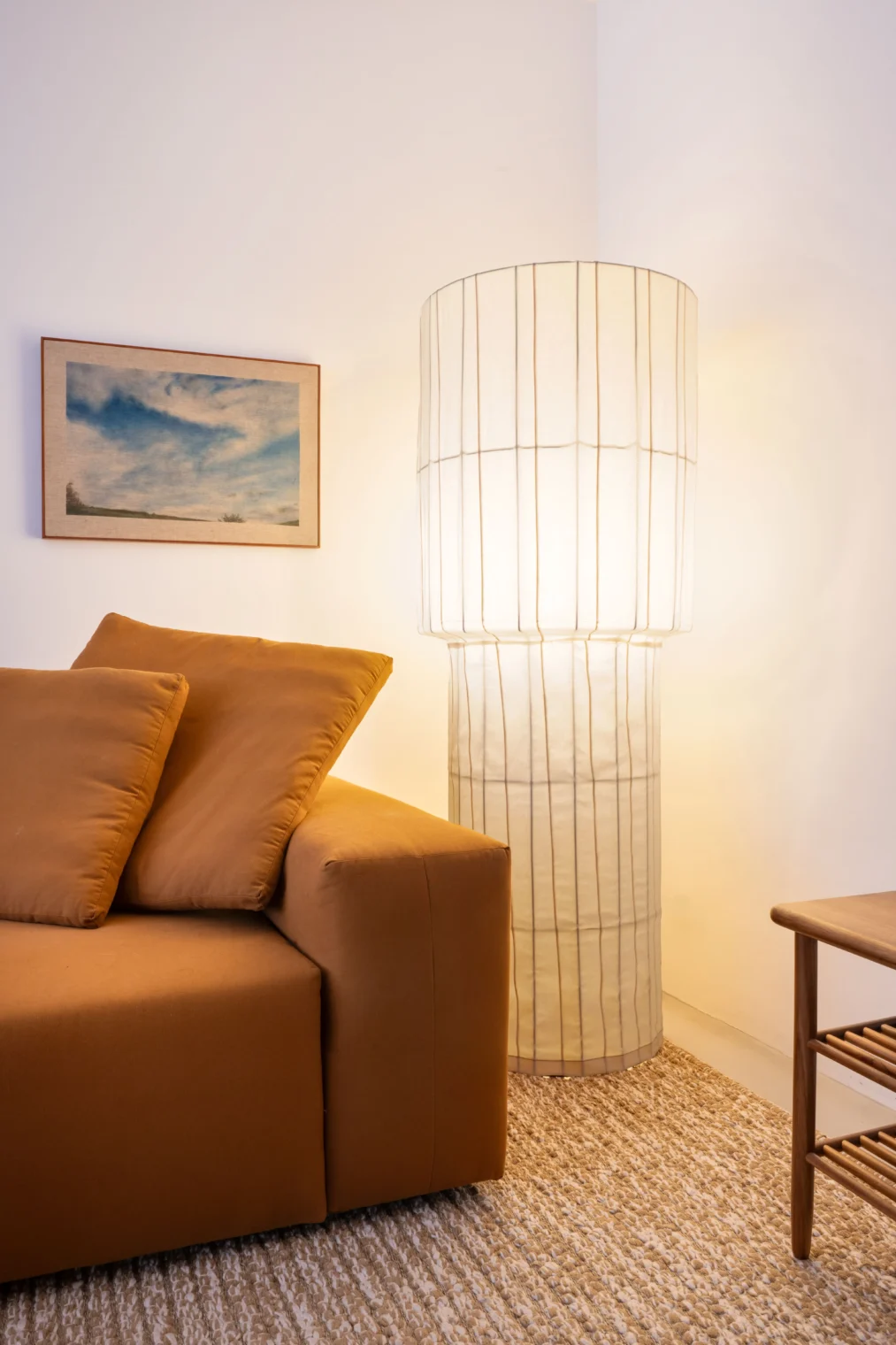 Detalhe de sala de estar com luminária de piso escultural de cúpula translúcida iluminada, sofá de tecido terracota, tapete de fibra natural e um quadro na parede branca.