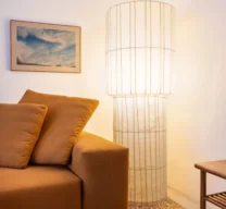 Detalhe de sala de estar com luminária de piso escultural de cúpula translúcida iluminada, sofá de tecido terracota, tapete de fibra natural e um quadro na parede branca.