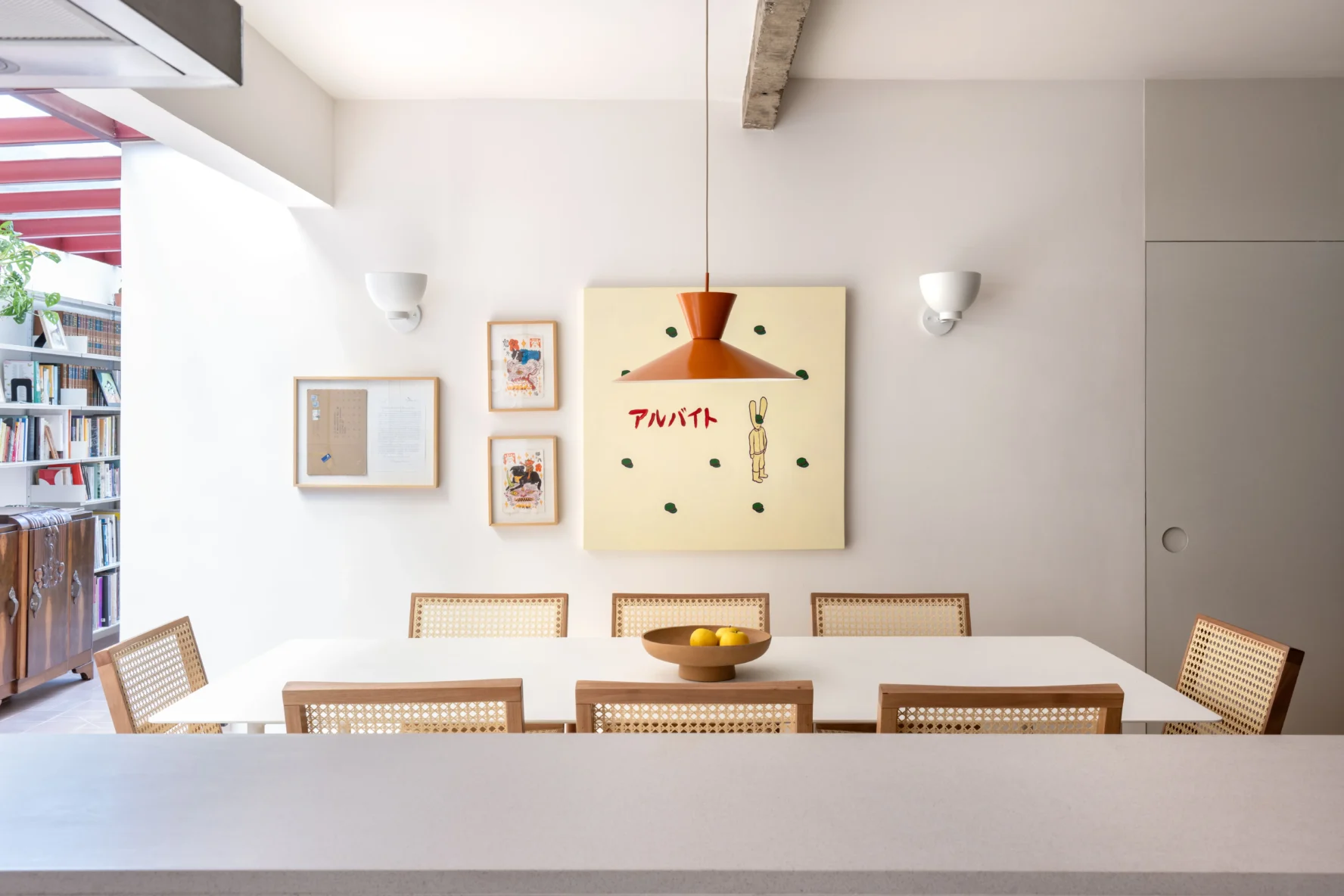 Vista da sala de jantar a partir da bancada, apresentando mesa branca, cadeiras de madeira com encosto em palhinha, pendente laranja e composição de quadros na parede.