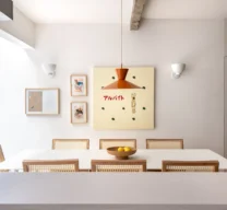 Vista da sala de jantar a partir da bancada, apresentando mesa branca, cadeiras de madeira com encosto em palhinha, pendente laranja e composição de quadros na parede.