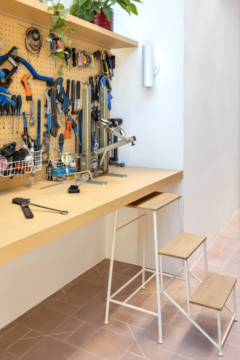 Detalhe de bancada de trabalho amarela clara com painel perfurado organizando diversas ferramentas, acompanhada por uma banqueta-escada metálica branca sobre piso de ladrilho geométrico rosa.