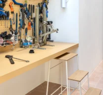 Detalhe de bancada de trabalho amarela clara com painel perfurado organizando diversas ferramentas, acompanhada por uma banqueta-escada metálica branca sobre piso de ladrilho geométrico rosa.