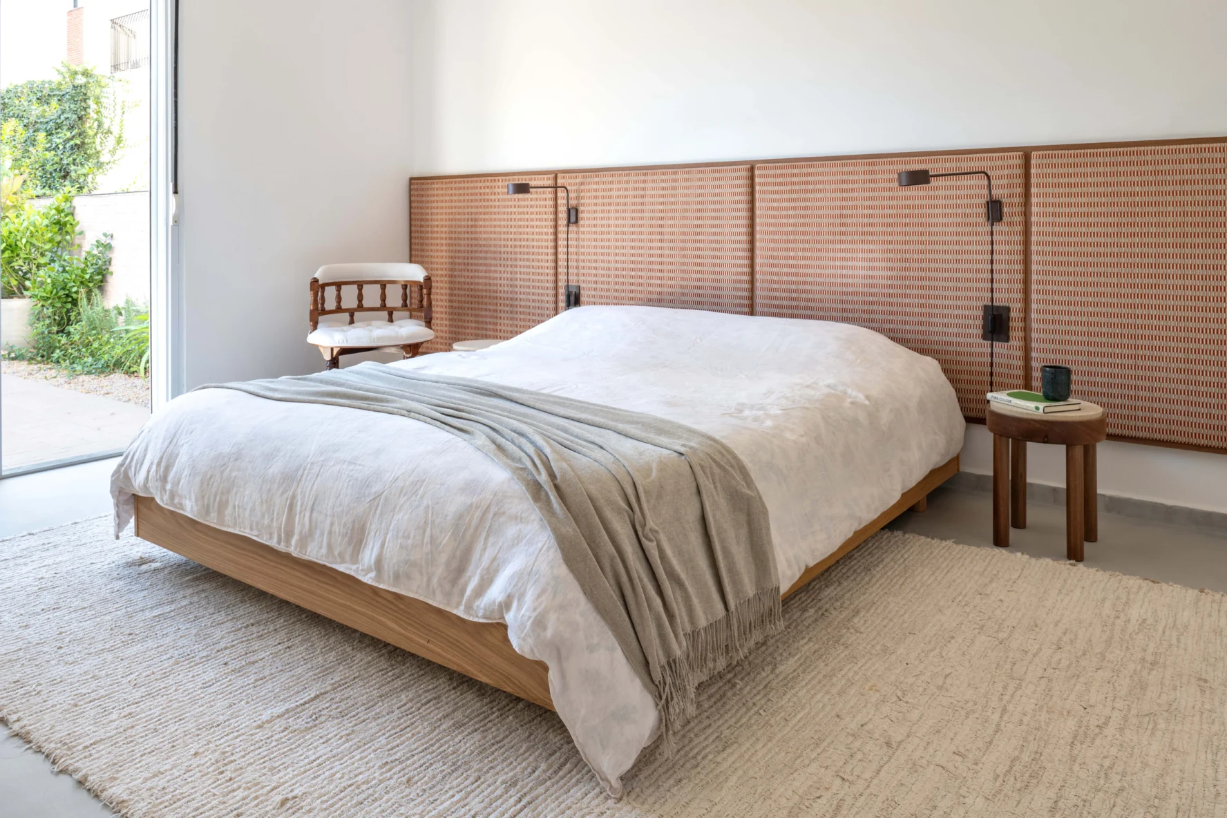 Projeto de quarto com cama de casal com base de madeira, grande painel de cabeceira em trama natural, tapete texturizado claro, arandelas e porta de vidro com vista para o jardim.