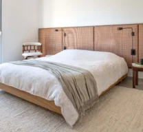 Projeto de quarto com cama de casal com base de madeira, grande painel de cabeceira em trama natural, tapete texturizado claro, arandelas e porta de vidro com vista para o jardim.