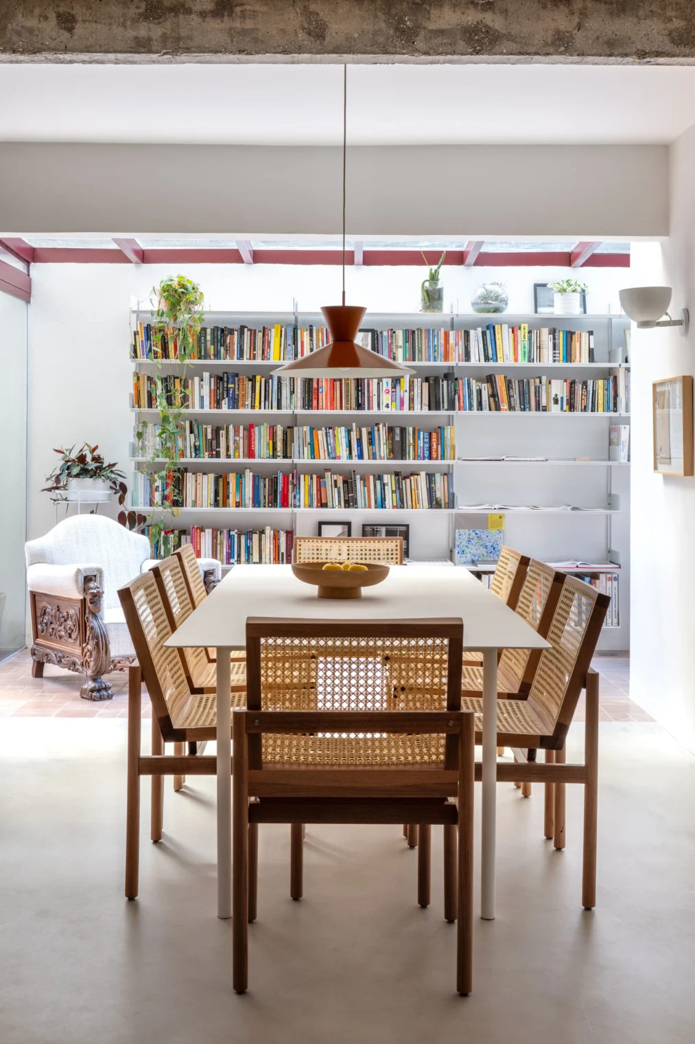 Vista frontal de uma sala de jantar com mesa branca, cadeiras de madeira e palhinha indiana, e pendente laranja. Ao fundo, uma área iluminada por teto de vidro com uma grande estante cheia de livros.