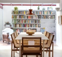 Vista frontal de uma sala de jantar com mesa branca, cadeiras de madeira e palhinha indiana, e pendente laranja. Ao fundo, uma área iluminada por teto de vidro com uma grande estante cheia de livros.