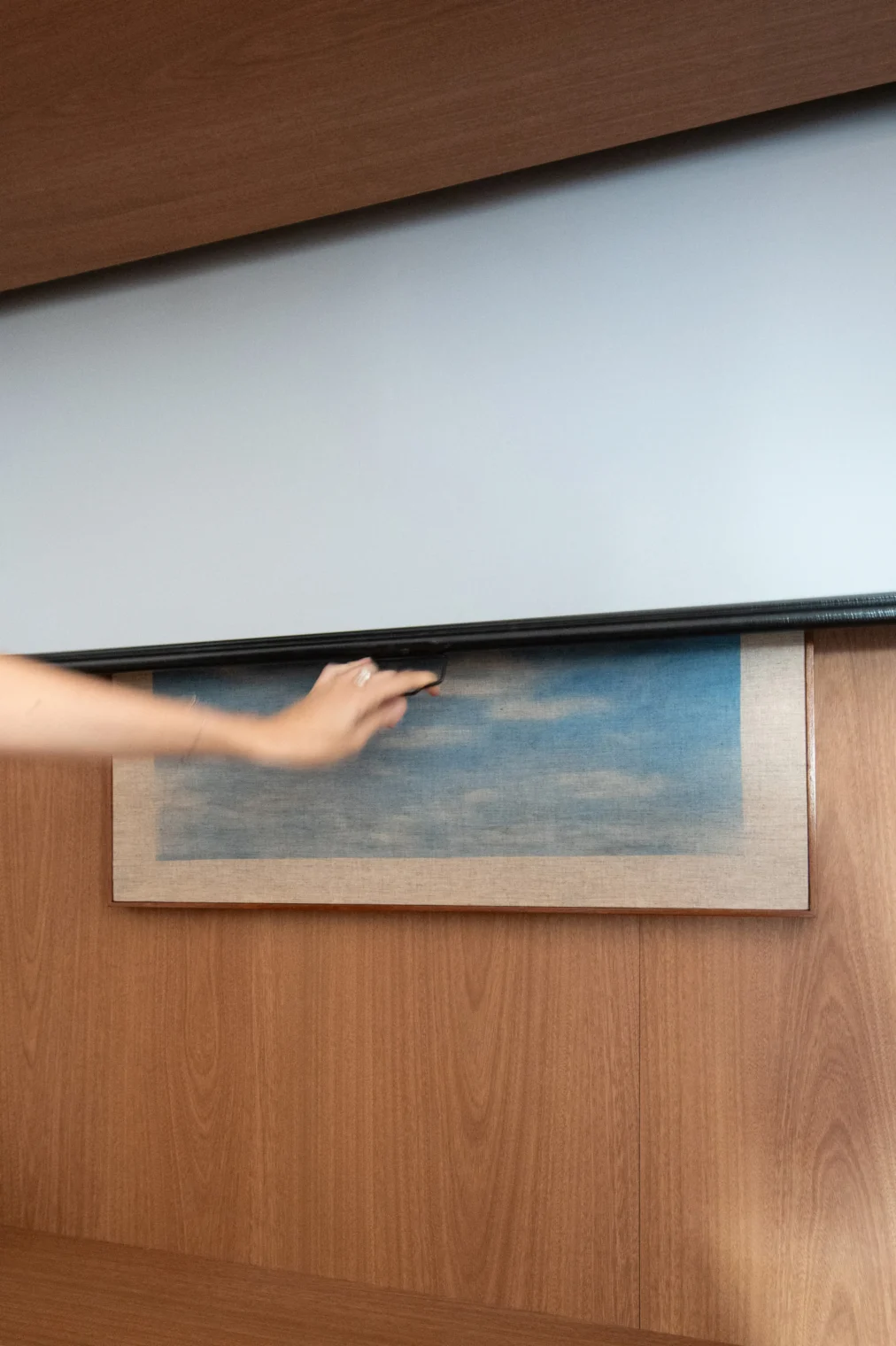 Detalhe de mão puxando uma tela retrátil de projeção embutida no nicho superior de marcenaria em madeira, cobrindo um quadro na parede quando puxada para baixo.