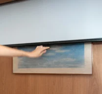 Detalhe de mão puxando uma tela retrátil de projeção embutida no nicho superior de marcenaria em madeira, cobrindo um quadro na parede quando puxada para baixo.