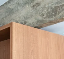 Foto em detalhe do encontro entre o topo de um móvel de marcenaria em madeira e uma viga estrutural de concreto aparente com acabamento bruto.