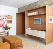Sala de estar com nicho aparador em marcenaria de madeira clara, viga aparente de concreto e sofá terracota sobre tapete claro.