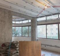 Fotografia de obra da sala em reforma no Apartamento Itacolomi com pilares de concreto aparente e perfis metálicos no teto, sobreposta por amostra de lâmina de madeira.