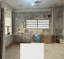 Fotografia de obra da cozinha em reforma no Apartamento Itacolomi com paredes descascadas e instalações expostas, sobreposta por amostra de pastilhas quadradas claras.
