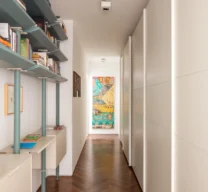 Corredor de circulação com estante em serralheria verde e creme à esquerda, armários com portas de correr lisas à direita, piso de taco em espinha de peixe e tela colorida ao fundo.
