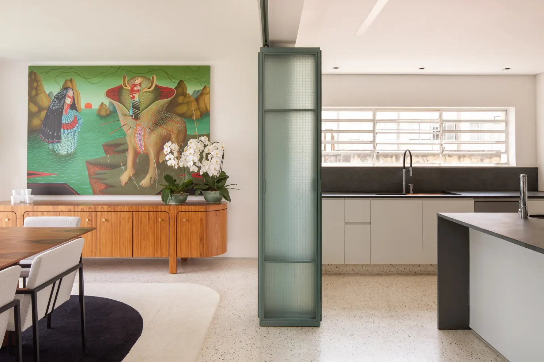 Transição entre sala de jantar e cozinha divididas por painel de correr em serralheria verde com vidro aramado, sobre piso em granilite claro.