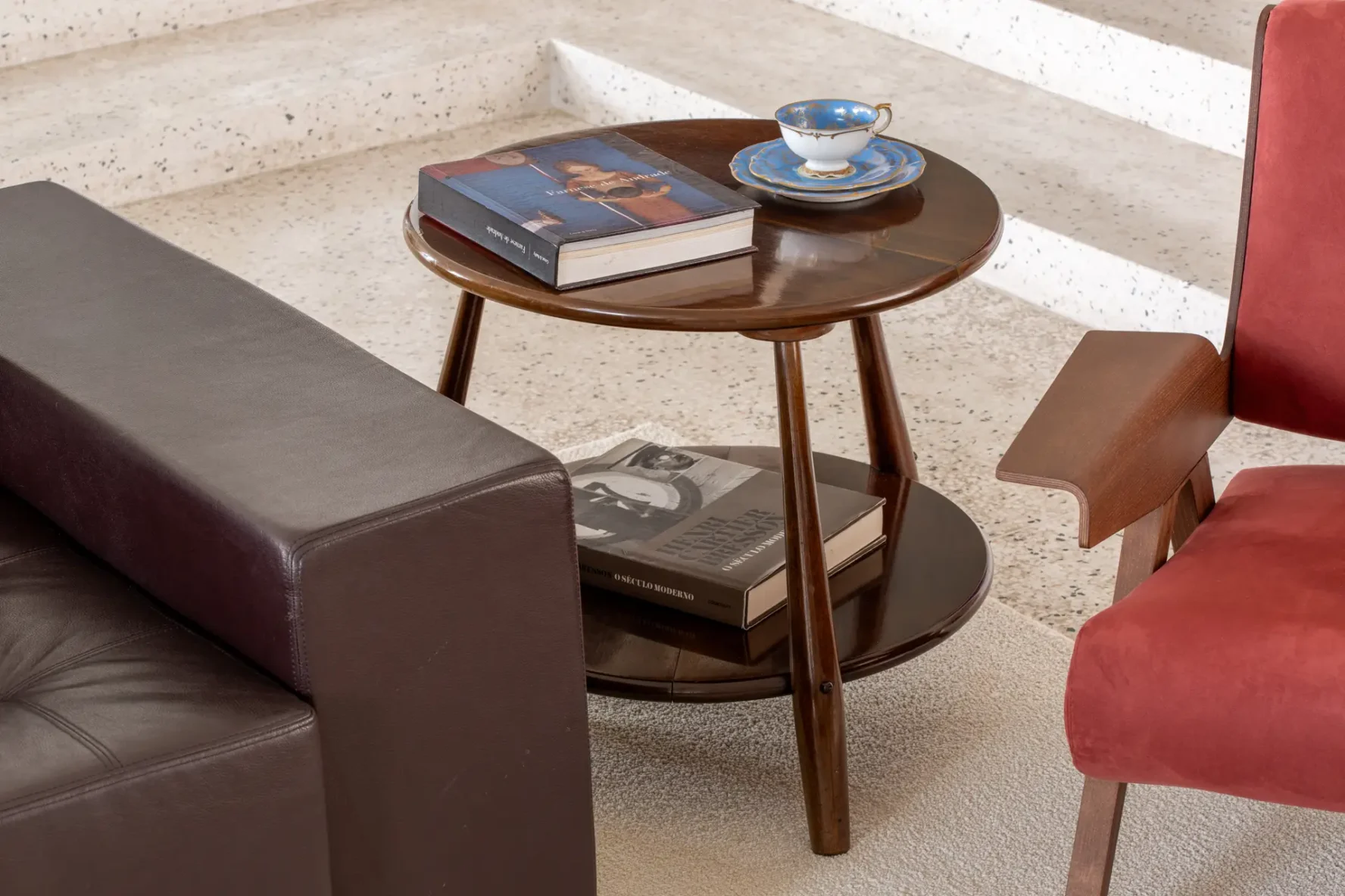 Detalhe de mesa lateral redonda de madeira de dois níveis entre um sofá de couro escuro e uma poltrona vermelha, apoiando livros de arte e xícara de porcelana azul.