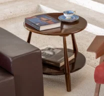 Detalhe de mesa lateral redonda de madeira de dois níveis entre um sofá de couro escuro e uma poltrona vermelha, apoiando livros de arte e xícara de porcelana azul.