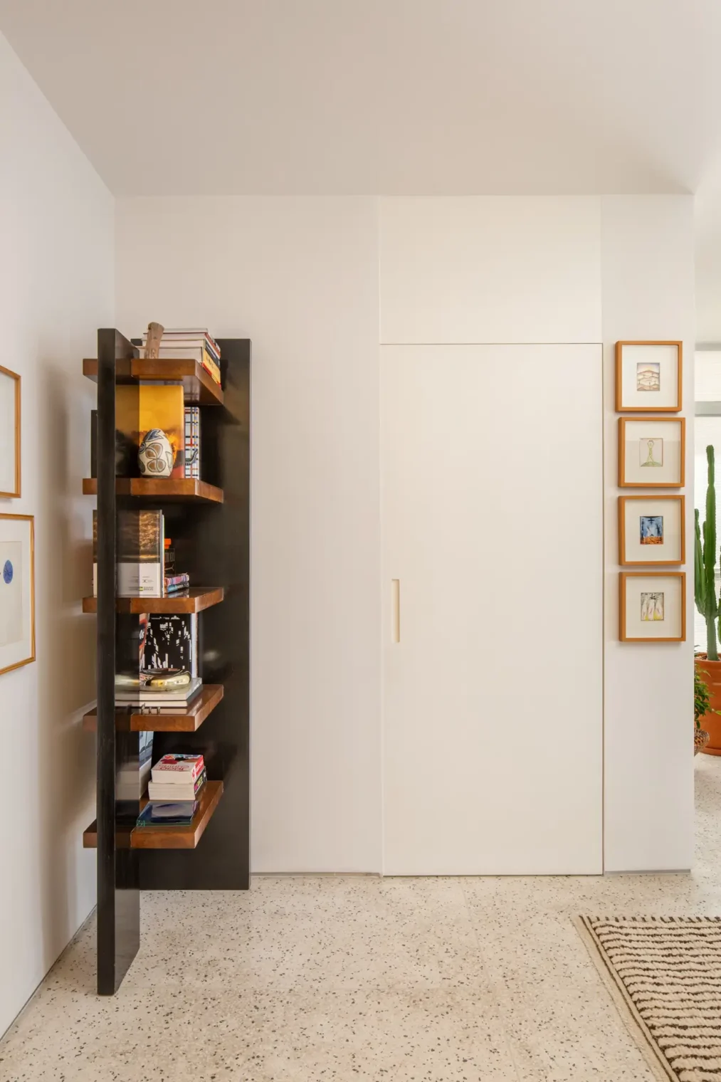 Hall de entrada com porta branca mimetizada no painel, estante vertical em marcenaria preta e madeira de um lado, e fileira de quadros pequenos do outro, sobre piso de granilite claro.