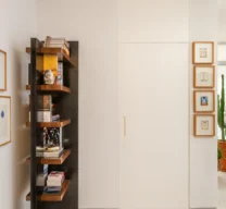 Hall de entrada com porta branca mimetizada no painel, estante vertical em marcenaria preta e madeira de um lado, e fileira de quadros pequenos do outro, sobre piso de granilite claro.