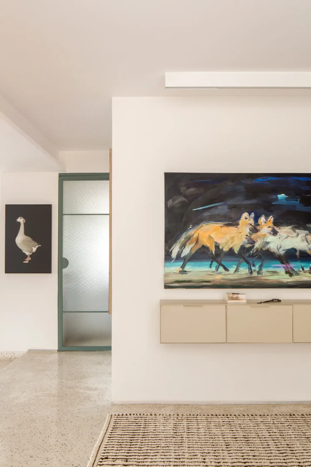 Hall com aparador suspenso creme em serralheria sob quadro de lobos-guará, porta em serralheria verde com vidro aramado ao fundo e piso em granilite claro com tapete listrado.
