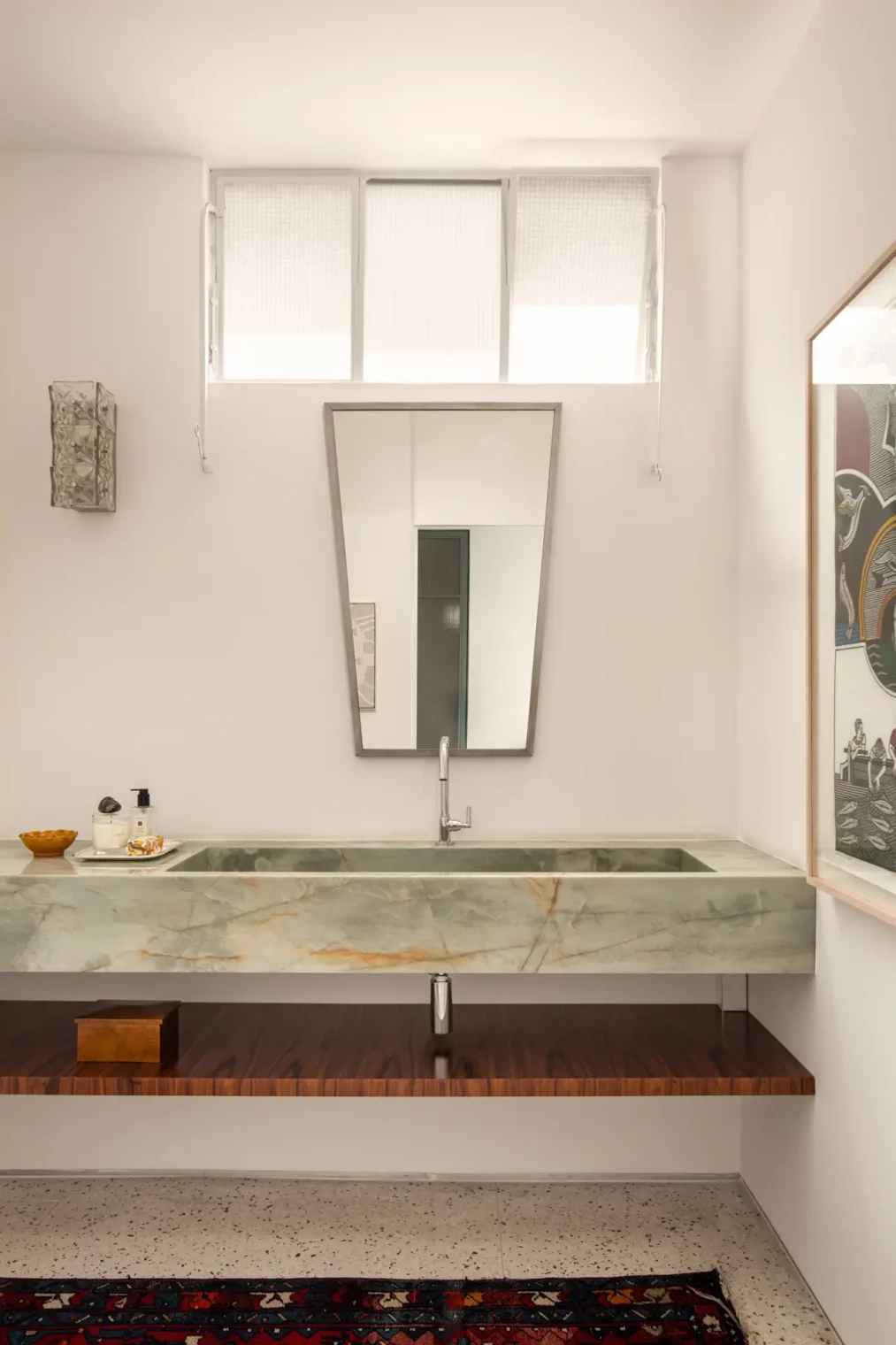 Lavabo com bancada de pedra natural esverdeada e cuba esculpida, espelho trapezoidal, prateleira inferior em madeira e piso de granilite claro.