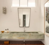 Lavabo com bancada de pedra natural esverdeada e cuba esculpida, espelho trapezoidal, prateleira inferior em madeira e piso de granilite claro.