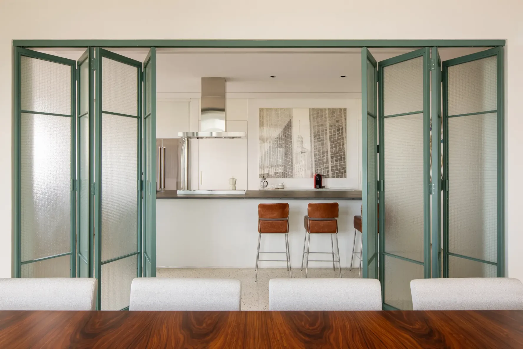 Porta camarão articulada em serralheria verde com vidro aramado aberta, integrando a sala de jantar à cozinha com ilha e banquetas de couro.