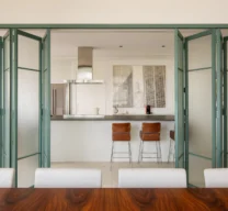Porta camarão articulada em serralheria verde com vidro aramado aberta, integrando a sala de jantar à cozinha com ilha e banquetas de couro.