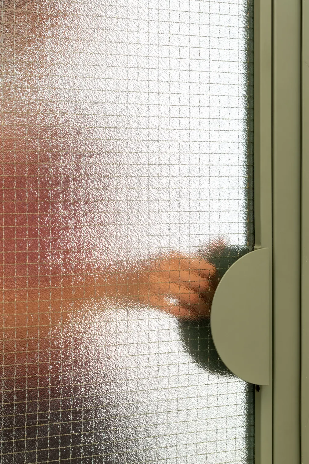 Detalhe de porta em serralheria verde com vidro aramado e puxador semicircular de chapa metálica, mostrando silhueta de mão do outro lado.