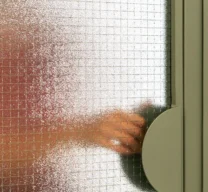 Detalhe de porta em serralheria verde com vidro aramado e puxador semicircular de chapa metálica, mostrando silhueta de mão do outro lado.