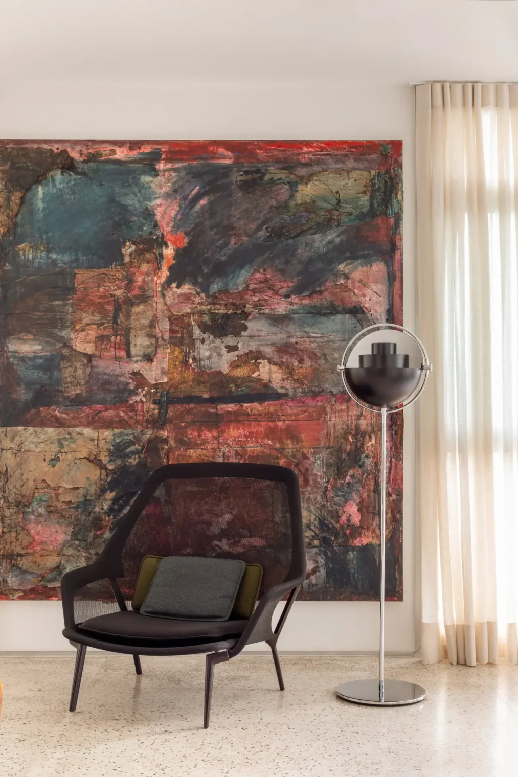 Cantinho com poltrona preta em tela, luminária de piso metálica esférica e grande quadro abstrato em tons de vermelho e escuros sobre piso de granilite claro.