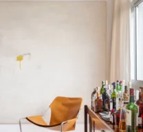 Cantinho de estar com poltrona Paulistano em couro caramelo, buffet em madeira servindo de bar e grande tela conceitual branca na parede sobre piso de granilite claro.